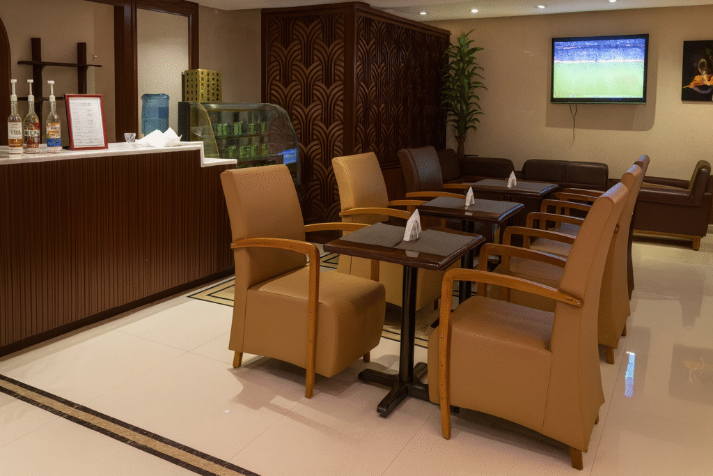 A modern indoor café-style lounge area with beige leather chairs, dark wooden tables, and a counter displaying various drink bottles. A decorative wooden partition, a television mounted on the wall, and indoor plants complete the cozy setting.